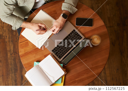 Top aerial view of male hands taking notes, writing in notebook, man sitting at round desk in cafe, working on laptop, studying Top aerial view of male hands taking notes, writing in notebook, man sitting at round desk in cafe, working on laptop, studying 112567225