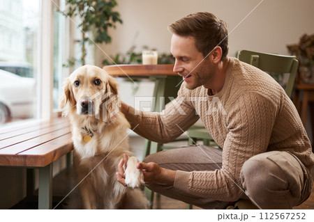 Portrait of young handsome man with his dog, sitting in cafe, golden retriever gives paw and looks at his owner 112567252