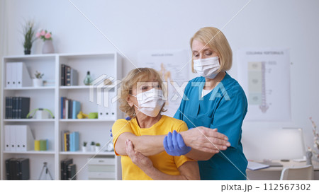 A woman therapist in a face mask helps a patient stretch her arm, relaxing their muscles 112567302