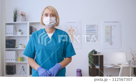 A woman doctor in a face mask dons medical gloves, preparing for a check-up or procedure A woman doctor in a face mask dons medical gloves, preparing for a check-up or procedure 112567307