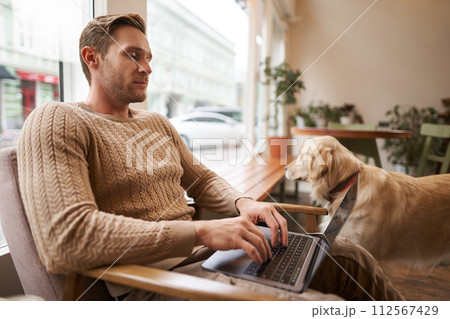 Handsome young man working in cafe with a dog, sitting on chair and using laptop, petting his golden retriever in animal-friendly co-working space Handsome young man working in cafe with a dog, sitting on chair and using laptop, petting his golden retriever in animal-friendly co-working space 112567429