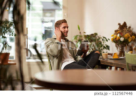 Portrait of handsome smiling young man, sitting in coffee shop, using digital tablet, video chats with someone from a cafe, wearing wireless headphones 112567446