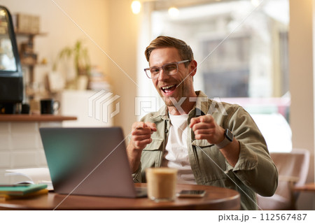 Happy young man in glasses, freelancer sitting in cafe with laptop, drinking coffee, pointing fingers at camera and smiling cheerfully 112567487