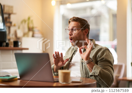 Portrait of man giving lessons online, shows okay, ok hand sign at laptop camera, talking to someone, having a meeting video chat, sitting in cafe 112567589