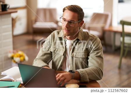 Portrait of young concentrated man in glasses, working from coffee shop, studying, looking thoughtful while completing task on laptop, making notes in notebook 112567602