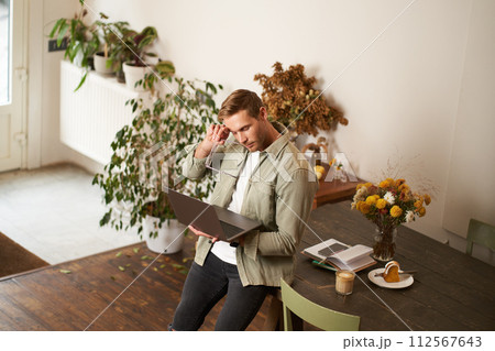 Image of young man, successful businessman sitting on table, looking at laptop, working in an office, looking concentrated on the project, doing job task, drinking coffee 112567643