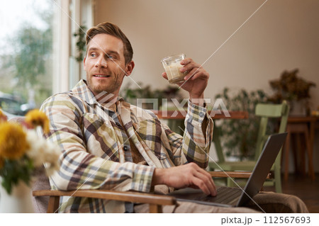 Portrait of young freelancer, a man in casual clothes, sitting in cafe and drinking coffee, working remotely on laptop, digital nomad doing his project outdoors in co-working space Portrait of young freelancer, a man in casual clothes, sitting in cafe and drinking coffee, working remotely on laptop, digital nomad doing his project outdoors in co-working space 112567693