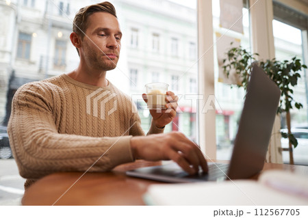 Portrait of handsome young man sitting in cafe with laptop. Digital nomad, freelancer working outdoors from coffee shop, typing something 112567705