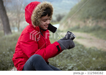Teen boy traveler taking a break, having a hot tea drink, sitting on forest log while contemplating the mountain scenery 112568162