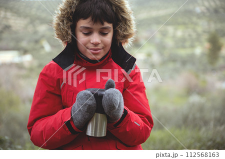 Authentic portrait of a smiling handsome traveler boy holding a thermos mug with hot drink, over nature background 112568163