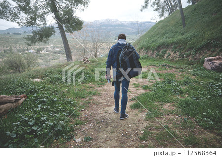 Rear view of a backpacked male tourist walking on the forest foot path in early spring nature, discovering mountains 112568364