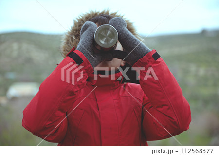 Teenager boy in red parka with fluffy further hood, drinking from steel travel thermos mug against mountains background 112568377
