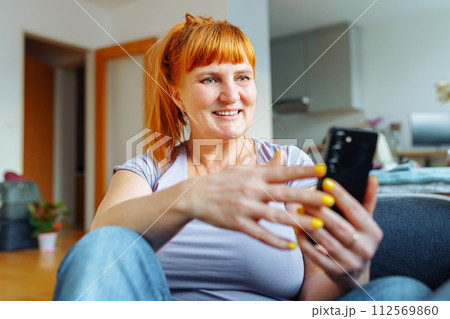 portrait red-haired woman using mobile phone while sitting on sofa at home in living room 112569860