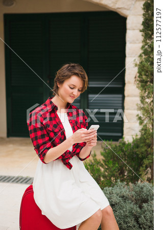 Young woman in a red shirt with a suitcase and phone on the city street.. 112577197