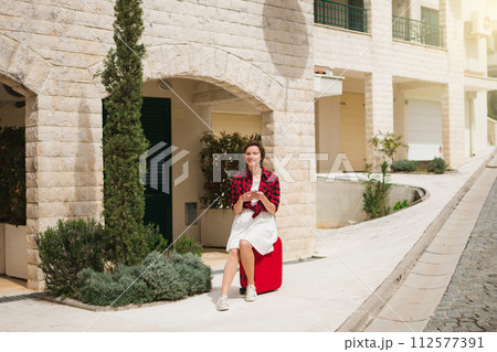 Young woman in a red shirt with a suitcase and phone on the city street.. 112577391