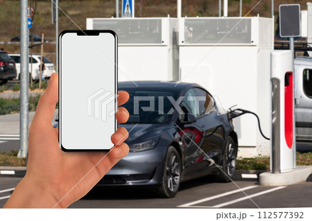 A hand with a phone on the background of a charging station for electric cars A hand with a phone on the background of a charging station for electric cars 112577392