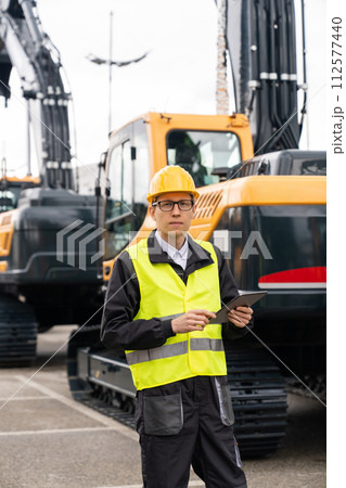 Engineer in a helmet with a digital tablet stands next to construction excavators.. 112577440