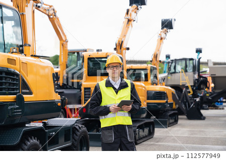 Engineer in a helmet with a digital tablet stands next to construction excavators.. Engineer in a helmet with a digital tablet stands next to construction excavators.. 112577949