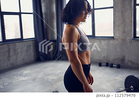 Side view portrait of concentrated young athlete woman in sportswear resting between workouts stands near barbell on floor Side view portrait of concentrated young athlete woman in sportswear resting between workouts stands near barbell on floor 112579470