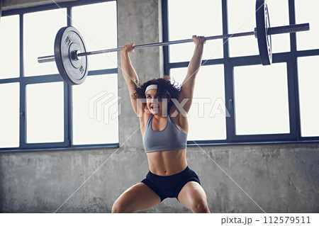 Side view of focused young strong woman in sportswear doing squat technique with barbell overhead in motion at gym. 112579511
