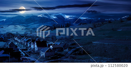 alpine countryside ant night. village in the valley and distant mountain range with snow capped tops in full moon light 112580020
