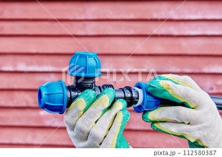 Plastic pipe tee connector fitting with compression clamp joint in hands of plumber on background of red wooden wall. Plastic pipe tee connector fitting with compression clamp joint in hands of plumber on background of red wooden wall. 112580387
