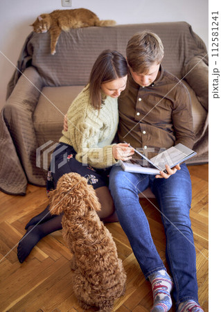 young couple looking at their wedding photo album by the bed in living room, cockapoo running around 112581241