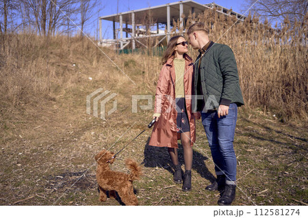 a young stylish couple walking with their cockapoo dog near the river, an unfinished parking lot in the background 112581274