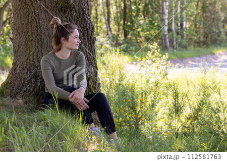 Joy in Nature's Lap: Smiling Woman Seated Under Tree 112581763
