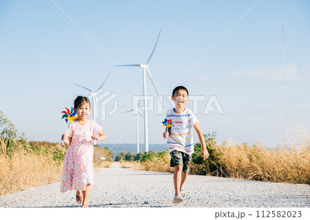 Running children boy and girl hold pinwheels near windmills. Clean energy visuals kids play joyfully embracing sustainability. Wind turbine family fun happiness. Industry future is windmill-generated. 112582023