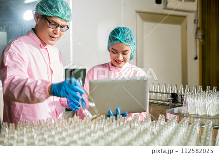 At the beverage factory two engineers meticulously check product bottles on the conveyor belt. Quality control and inspection are diligently managed by an engineer and inspector using a laptop. 112582025