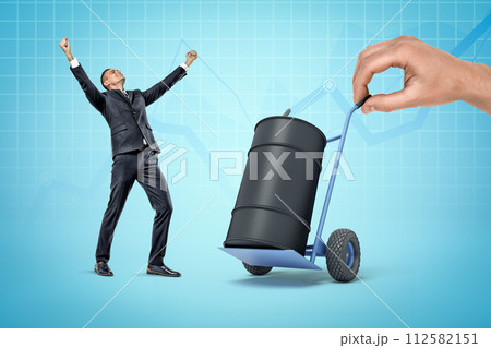 Very happy businessman standing near big black oil barrel on hand truck held by huge hand on blue background. 112582151