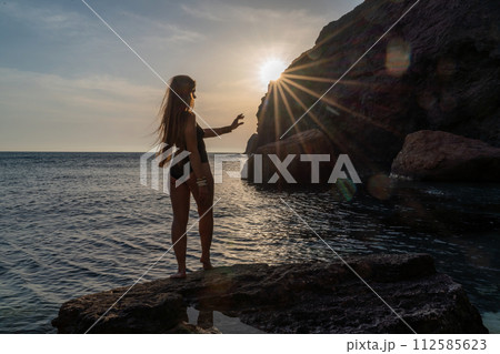 Woman tourist enjoying the sunset over the sea mountain landscape. Sits outdoors on a rock above the sea. She is wearing jeans and a blue hoodie. Healthy lifestyle, harmony and meditation Woman tourist enjoying the sunset over the sea mountain landscape. Sits outdoors on a rock above the sea. She is wearing jeans and a blue hoodie. Healthy lifestyle, harmony and meditation 112585623