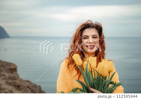 Portrait of a happy woman with hair flying in the wind against the backdrop of mountains and sea. Holding a bouquet of yellow tulips in her hands, wearing a yellow sweater 112585844