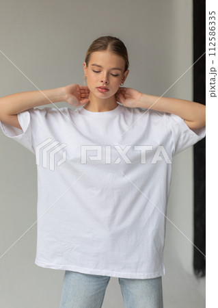 Beautiful blonde girl in a white oversized T-shirt and blue jeans posing on a gray background Beautiful blonde girl in a white oversized T-shirt and blue jeans posing on a gray background 112586335
