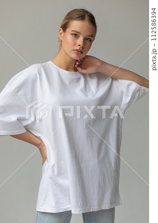 Beautiful blonde girl in a white oversized T-shirt and blue jeans posing on a gray background Beautiful blonde girl in a white oversized T-shirt and blue jeans posing on a gray background 112586394