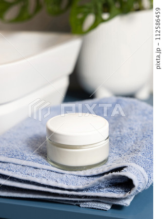 Cosmetic cream jar on blue towels near basin and green monstera in bath, lid mockup 112586459