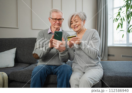 Senior couple man woman shopping online with smartphone paying with credit card. Old people buying on Internet enter credit card details at home indoors. Online shopping delivery service ecommerce Senior couple man woman shopping online with smartphone paying with credit card. Old people buying on Internet enter credit card details at home indoors. Online shopping delivery service ecommerce 112586940