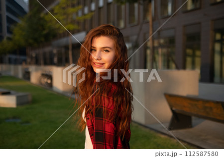 A young smiling red haired woman in a plaid shirt stands against the background of a business center A young smiling red haired woman in a plaid shirt stands against the background of a business center 112587580