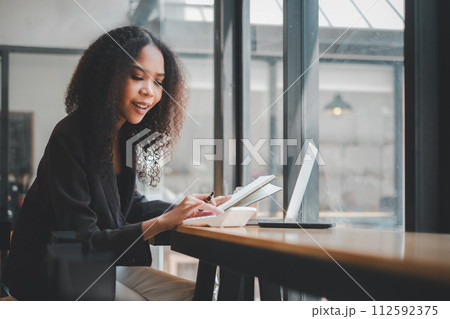 Portrait of attractive hardworking businesswoman with Afro hairstyle busy doing paperwork at office desk, working through finances, using calculator and making notes in her notebook with pen Portrait of attractive hardworking businesswoman with Afro hairstyle busy doing paperwork at office desk, working through finances, using calculator and making notes in her notebook with pen 112592375