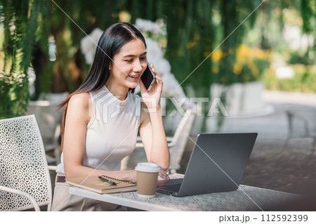 Smiling young woman having a pleasant conversation on her mobile phone while sitting with her laptop at an outdoor table. 112592399