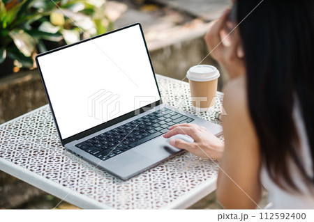 Over the shoulder shot of a woman working on a laptop with a blank screen, suitable for mock-ups, with a coffee cup beside her. 112592400