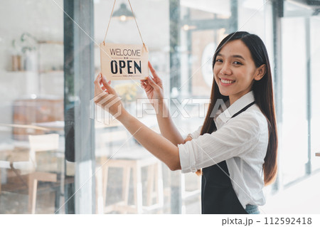 A dedicated cafe owner hangs an inviting "Welcome Open sign", signaling the start of the day in her cozy coffee shop, an emblem of small business hospitality and readiness. A dedicated cafe owner hangs an inviting "Welcome Open sign", signaling the start of the day in her cozy coffee shop, an emblem of small business hospitality and readiness. 112592418