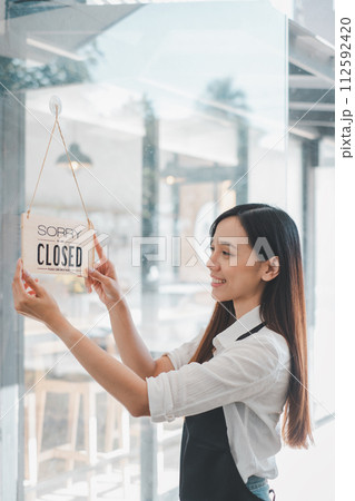 A cafe owner displays a "Sorry We Are Closed" sign with a content smile, marking the end of a fulfilling day of business in her welcoming establishment. A cafe owner displays a "Sorry We Are Closed" sign with a content smile, marking the end of a fulfilling day of business in her welcoming establishment. 112592420