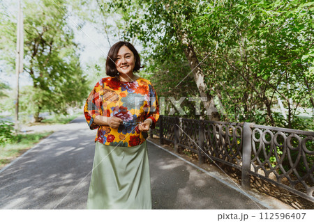 Asian woman with a joyful, open smile enjoys the tranquility of a summer day while walking along the sidewalk of the park Smiling middle aged woman walking, relaxing in park and radiating happiness Asian woman with a joyful, open smile enjoys the tranquility of a summer day while walking along the sidewalk of the park Smiling middle aged woman walking, relaxing in park and radiating happiness 112596407