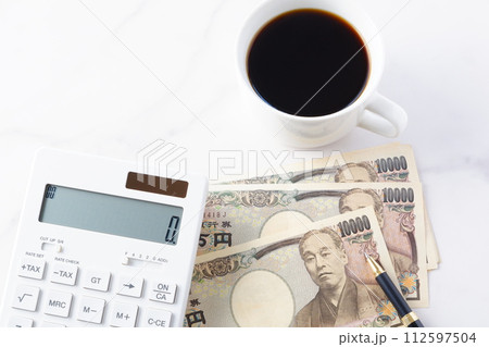 福沢諭吉の肖像の一万円紙幣と電卓、ペン、淹れたてのコーヒーが入ったコーヒーカップ 112597504