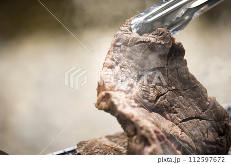 バーベキューで分厚いステーキ肉をトングで持ち上げる女性手元 バーベキューで分厚いステーキ肉をトングで持ち上げる女性手元 112597672