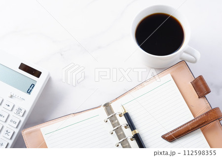 淹れたてのコーヒーを飲みながら、電卓とリング式のシステム手帳でデスクワークをしているイメージ 112598355