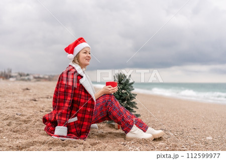 Sea Lady in Santa hat plaid shirt with a red mug in her hands enjoys beach with Christmas tree. Coastal area. Christmas, New Year holidays concep 112599977