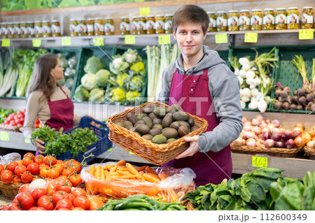 Young male seller holding avocados standing by counter in grocery market 112600349